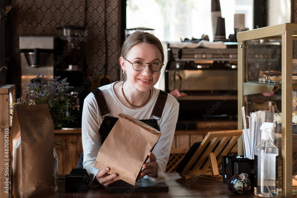 Fototapeta premium Young smiling woman (salesperson in bakery) holding craft paper packing bag for taking away food in coffee shop