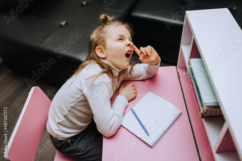 Tablou pe pânză Funny, cheerful, lazy red-haired girl, child, schoolgirl sits at a wooden pink desk at school and picks her nose