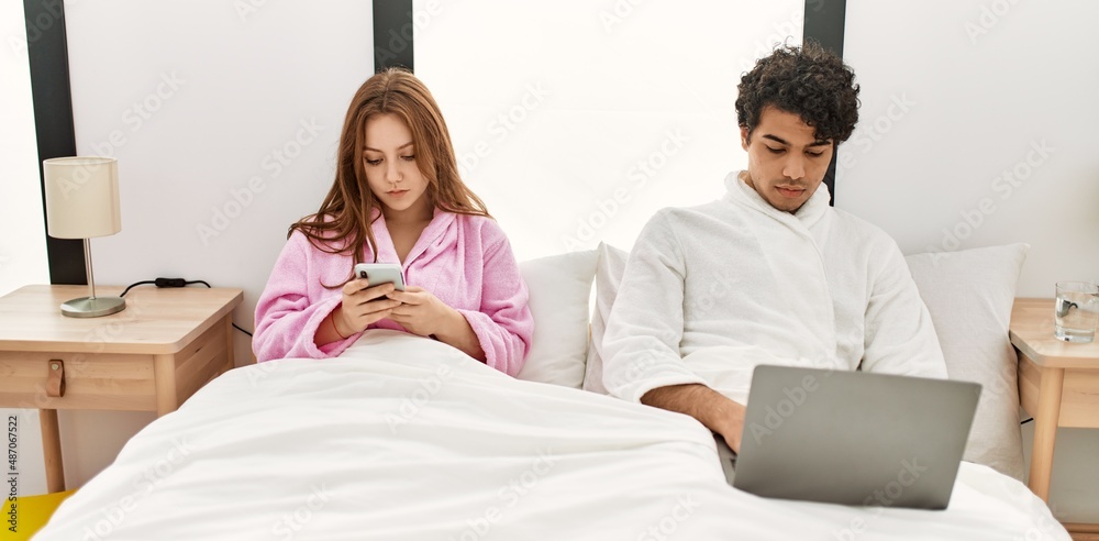 Fototapeta premium Young couple with serious expression using laptop and smartphone at bedroom.