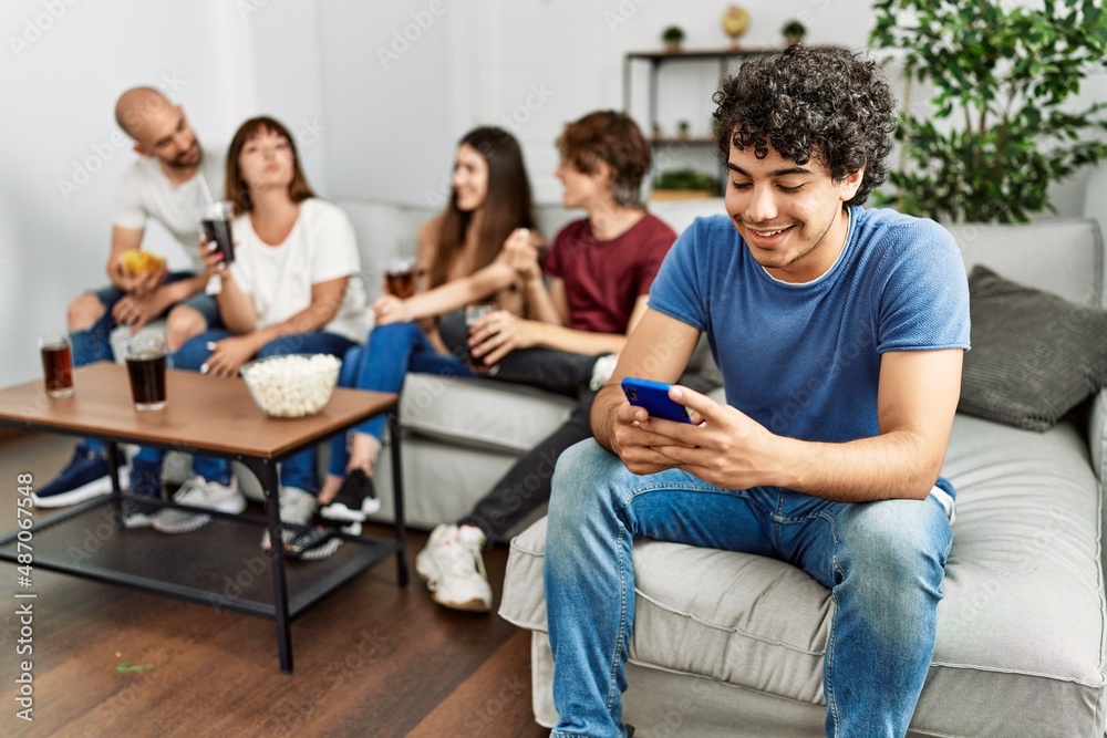 Group of young friends having party sitting on the sofa at home. Man smiling happy using smartphone at home. Group of young friends having party sitting on the sofa at home. Man smiling happy using smartphone at home.