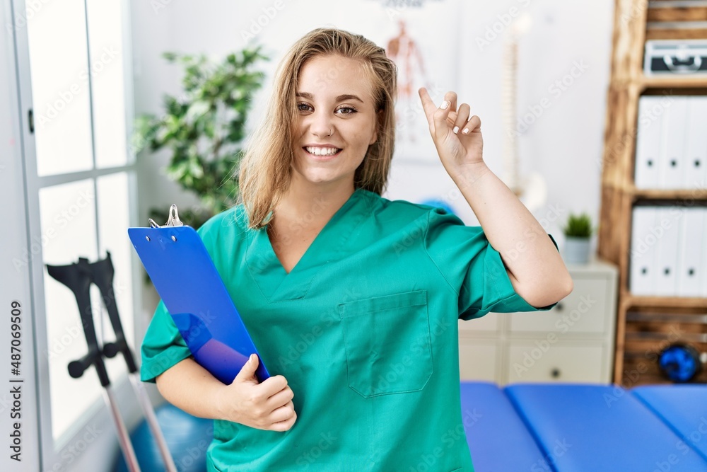Young caucasian woman working at pain recovery clinic smiling amazed and surprised and pointing up with fingers and raised arms. Young caucasian woman working at pain recovery clinic smiling amazed and surprised and pointing up with fingers and raised arms.