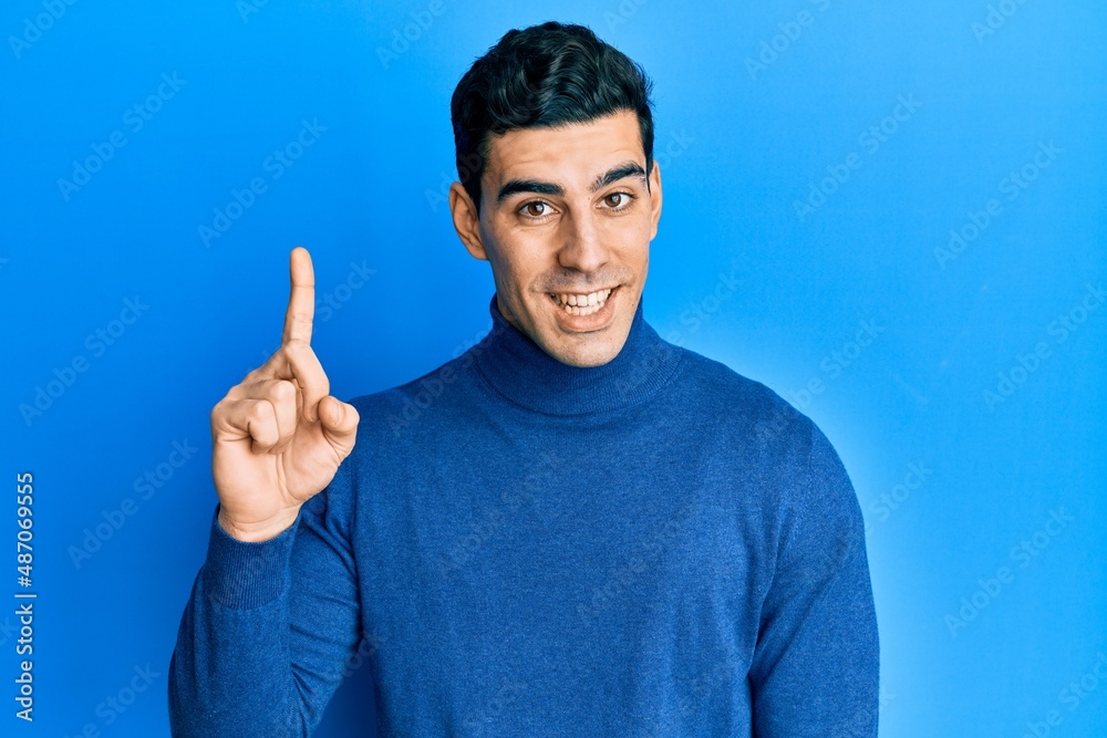 Naklejka premium Handsome hispanic man wearing turtleneck sweater smiling with an idea or question pointing finger up with happy face, number one