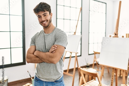 Young hispanic artist man smiling happy standing with arms crossed gesture at art studio.