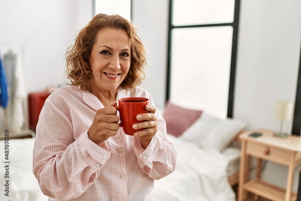 Middle age caucasian woman drinking coffee standing at bedroom.