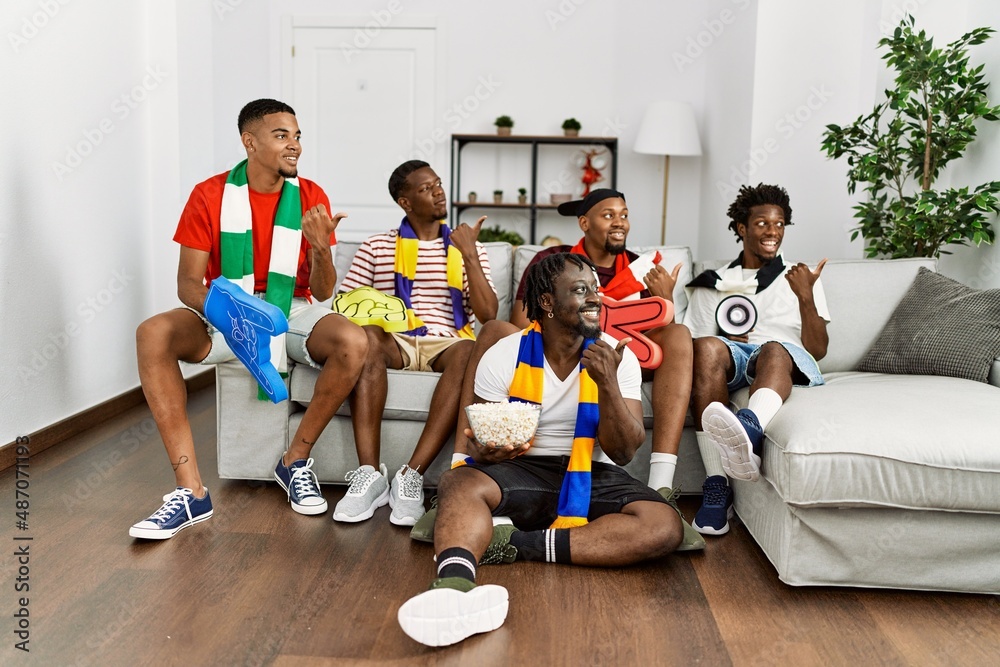 Fototapeta premium Young african men wearing team scarf cheering football game pointing thumb up to the side smiling happy with open mouth
