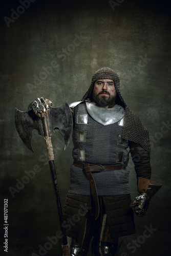 Φωτογραφία Vintage portrait of brutal seriuos man, medieval warrior or knight with wounded face in chain mail holding ax isolated over dark background