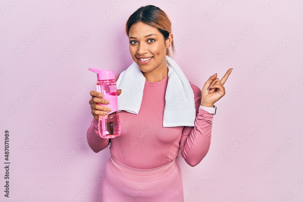 Beautiful hispanic woman wearing sportswear holding water bottle smiling happy pointing with hand and finger to the side Beautiful hispanic woman wearing sportswear holding water bottle smiling happy pointing with hand and finger to the side