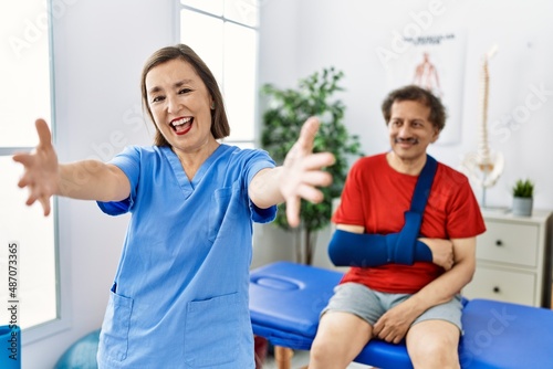 Middle age doctor woman with patient with arm injury at rehabilitation clinic looking at the camera smiling with open arms for hug. cheerful expression embracing happiness.