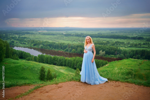 pregnant woman in blue dress, green hills, river and sky. High quality photo