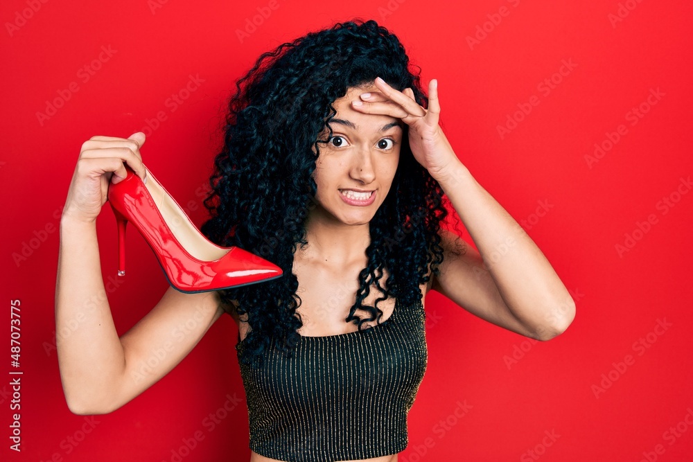 Young hispanic woman with curly hair holding red high heel shoes stressed and frustrated with hand on head, surprised and angry face Young hispanic woman with curly hair holding red high heel shoes stressed and frustrated with hand on head, surprised and angry face