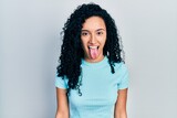 Young hispanic woman with curly hair wearing casual blue t shirt sticking tongue out happy with funny expression. emotion concept.