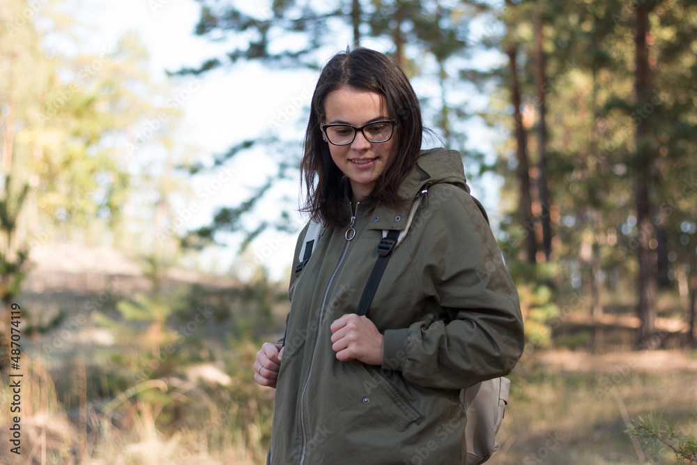 beautiful girl with dark long hair in glasses looks away while standing in the forest with a backpack beautiful girl with dark long hair in glasses looks away while standing in the forest with a backpack