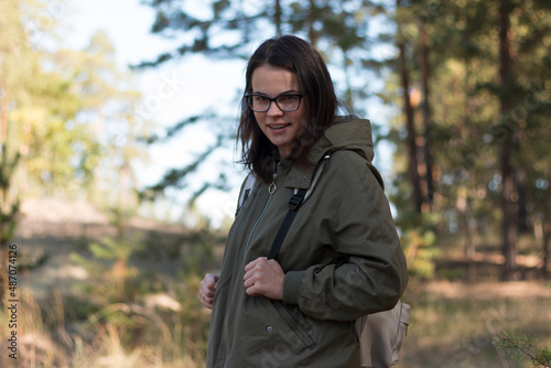 beautiful girl with dark long hair in glasses looks away while standing in the forest with a backpack © PeterPike