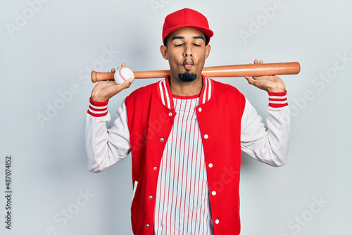 Young african american man playing baseball holding bat and ball making fish face with mouth and squinting eyes, crazy and comical. photo