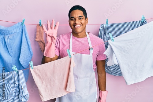 Young handsome hispanic man wearing cleaner apron holding clothes on clothesline showing and pointing up with fingers number four while smiling confident and happy.