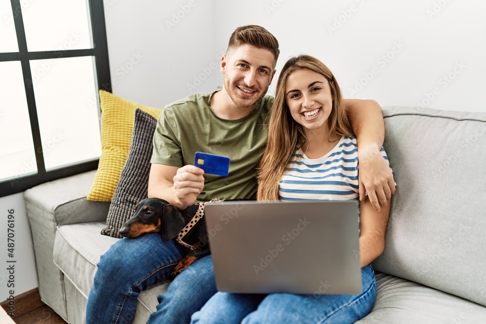 Young hispanic couple smiling happy using laptop and credit card sitting on the sofa  with dog at home.