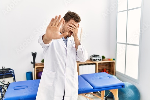Handsome young man working at pain recovery clinic covering eyes with hands and doing stop gesture with sad and fear expression. embarrassed and negative concept.