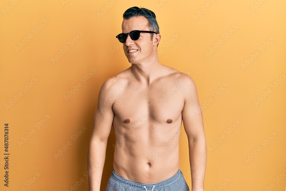Handsome young man wearing swimsuit and sunglasses looking to side, relax profile pose with natural face and confident smile.