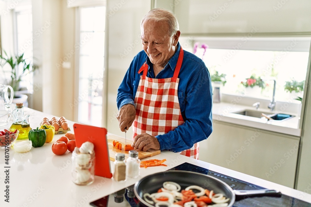 Senior man smiling confident cooking and watching online recipe at kitchen