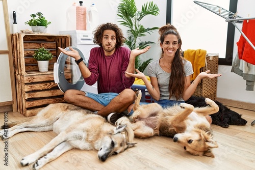 Young hispanic couple doing laundry with dogs clueless and confused expression with arms and hands raised. doubt concept.