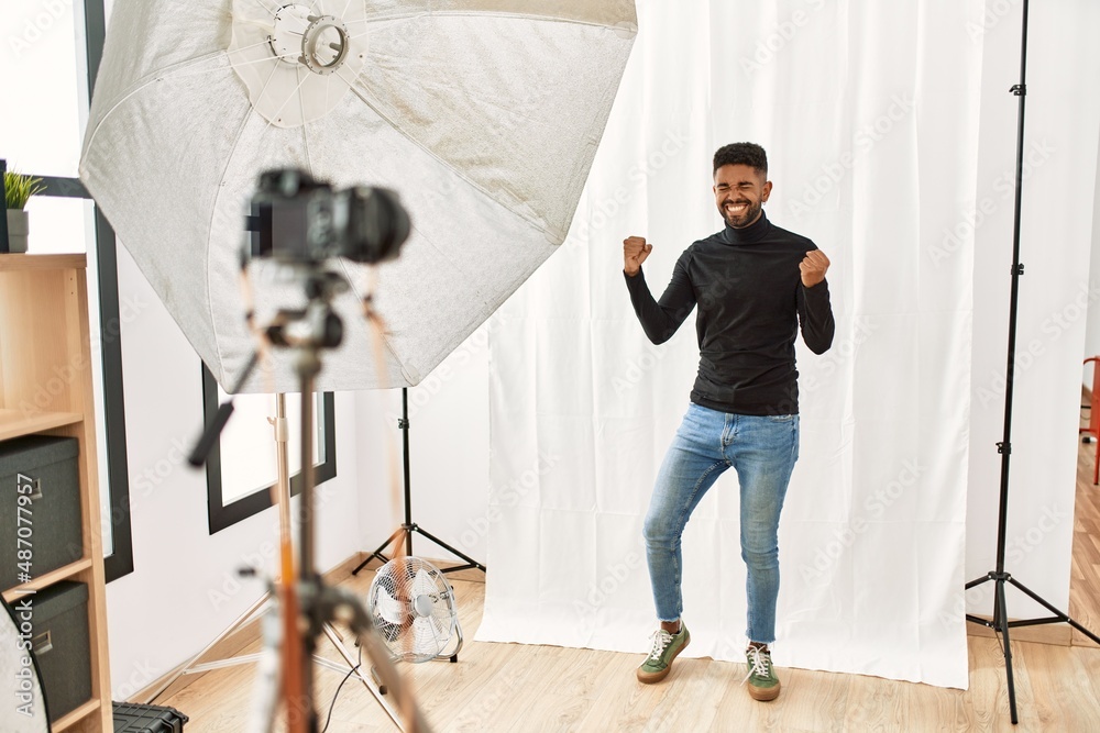 Young hispanic man with beard posing as model at photography studio very happy and excited doing winner gesture with arms raised, smiling and screaming for success. celebration concept.