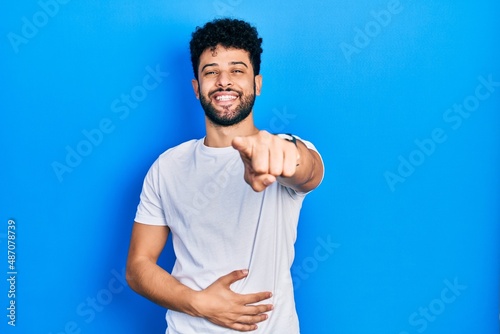 Young arab man with beard wearing casual white t shirt laughing at you, pointing finger to the camera with hand over body, shame expression