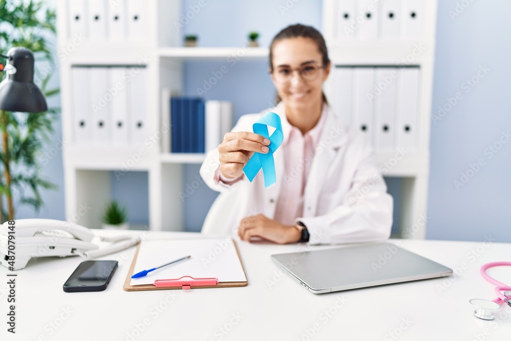 Young hispanic woman wearing doctor uniform holding blue ribbon at clinic