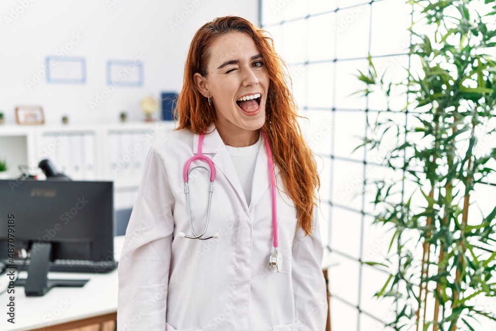 Young redhead woman wearing doctor uniform and stethoscope at the clinic winking looking at the camera with sexy expression, cheerful and happy face. Young redhead woman wearing doctor uniform and stethoscope at the clinic winking looking at the camera with sexy expression, cheerful and happy face.