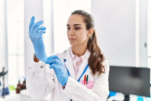Young hispanic woman wearing scientist uniform wearing gloves at laboratory