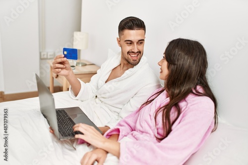 Young hispanic couple using laptop and credita card lying in bed at home.