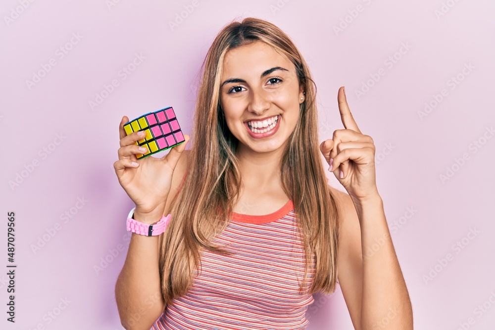 Beautiful hispanic woman holding colorful puzzle cube intelligence game smiling with an idea or question pointing finger with happy face, number one