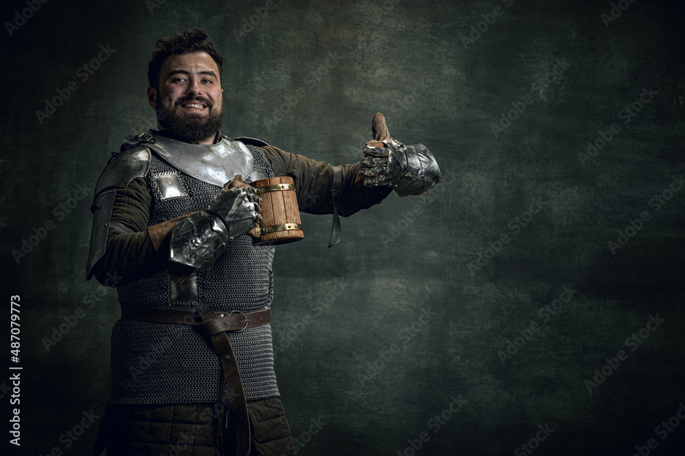 Naklejka premium Happy medieval warrior or knight with dirty wounded face holding big mug of beer isolated over dark vintage background. Comparison of eras, history