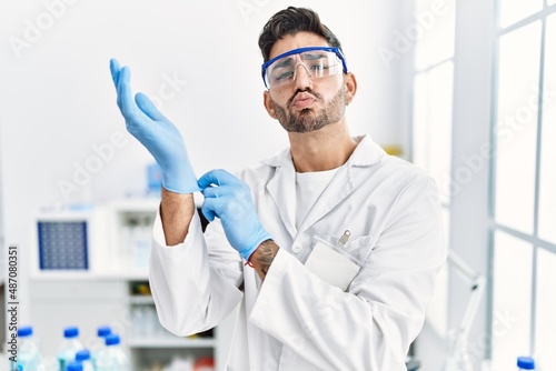 Young hispanic man working at scientist laboratory putting gloves on looking at the camera blowing a kiss being lovely and sexy. love expression.