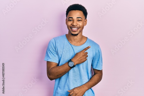 Young african american man wearing casual blue t shirt cheerful with a smile on face pointing with hand and finger up to the side with happy and natural expression