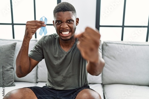 Young african american man holding blue ribbon sitting on the sofa at home annoyed and frustrated shouting with anger, yelling crazy with anger and hand raised © Krakenimages.com