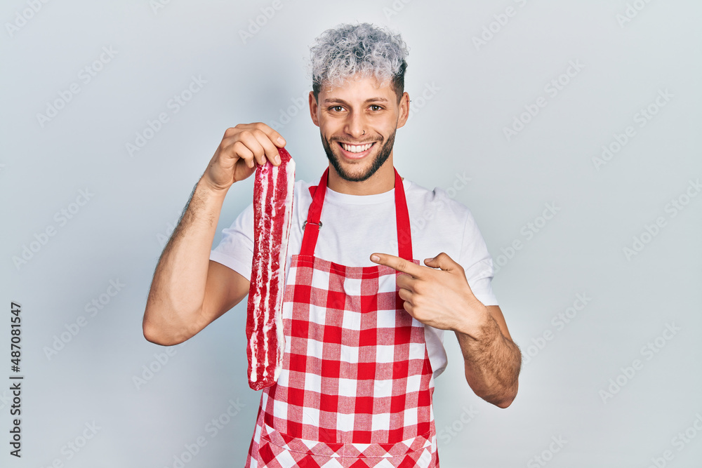 Young hispanic man with modern dyed hair wearing apron holding raw beef steak smiling happy pointing with hand and finger Young hispanic man with modern dyed hair wearing apron holding raw beef steak smiling happy pointing with hand and finger
