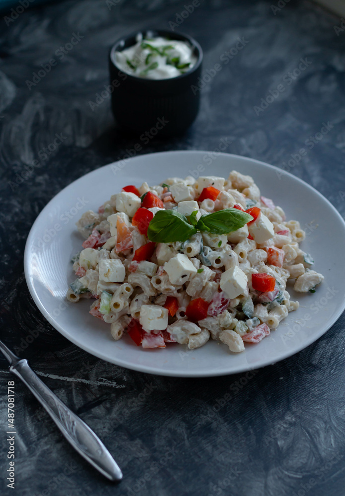 Pasta salad with red pepper, feta cheese and basil on a white plate, next to it there is a fork and a bowl with sauce Pasta salad with red pepper, feta cheese and basil on a white plate, next to it there is a fork and a bowl with sauce