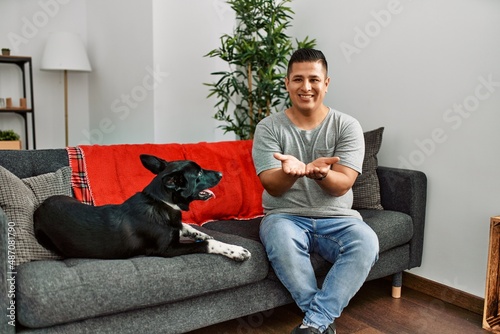 Young latin man and dog sitting on the sofa at home smiling with hands palms together receiving or giving gesture. hold and protection © Krakenimages.com