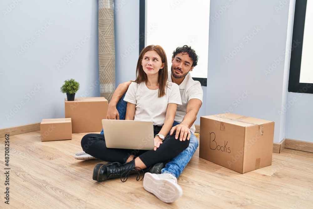 Young couple using laptop at new home smiling looking to the side and staring away thinking.