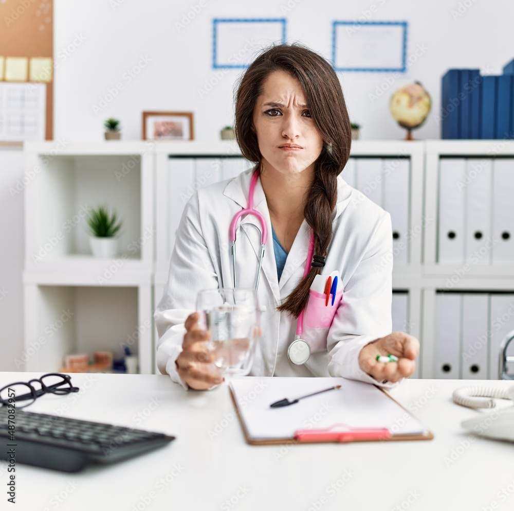 Young doctor woman holding glass of water and prescription pills skeptic and nervous, frowning upset because of problem. negative person. Young doctor woman holding glass of water and prescription pills skeptic and nervous, frowning upset because of problem. negative person.