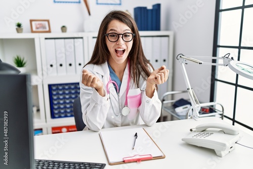 Young doctor woman wearing doctor uniform and stethoscope at the clinic celebrating surprised and amazed for success with arms raised and open eyes. winner concept.