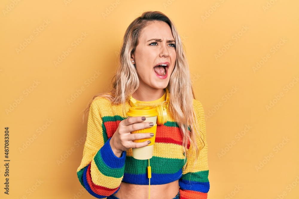 Beautiful young blonde woman drinking cup of coffee wearing headphones angry and mad screaming frustrated and furious, shouting with anger. rage and aggressive concept. Beautiful young blonde woman drinking cup of coffee wearing headphones angry and mad screaming frustrated and furious, shouting with anger. rage and aggressive concept.