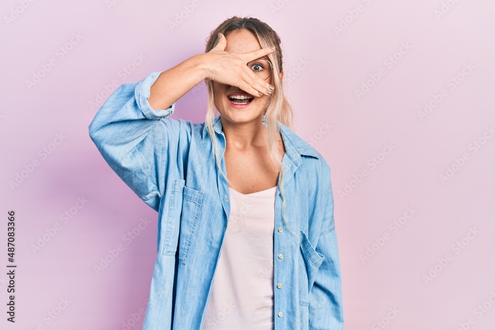 Beautiful young blonde woman wearing casual clothes and glasses peeking in shock covering face and eyes with hand, looking through fingers with embarrassed expression. Beautiful young blonde woman wearing casual clothes and glasses peeking in shock covering face and eyes with hand, looking through fingers with embarrassed expression.