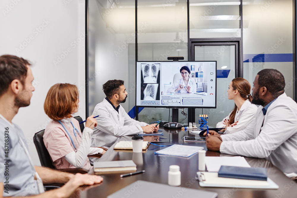 Large group of intercultural medical workers looking at their colleague on computer screen while watching online presentation Large group of intercultural medical workers looking at their colleague on computer screen while watching online presentation