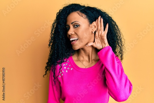 Middle age african american woman wearing casual clothes smiling with hand over ear listening and hearing to rumor or gossip. deafness concept.