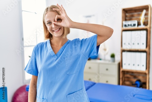 Young caucasian physiotherapist woman working at pain recovery clinic smiling happy doing ok sign with hand on eye looking through fingers