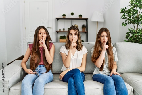 Group of three hispanic girls sitting on the sofa at home asking to be quiet with finger on lips. silence and secret concept.