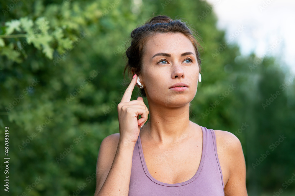 Close-up Portrait of beautiful young, pensive woman talking through bluetooth headphones while at sports jog. A serious girl, listens to music with, bluetooth wireless headphones. Close-up Portrait of beautiful young, pensive woman talking through bluetooth headphones while at sports jog. A serious girl, listens to music with, bluetooth wireless headphones.