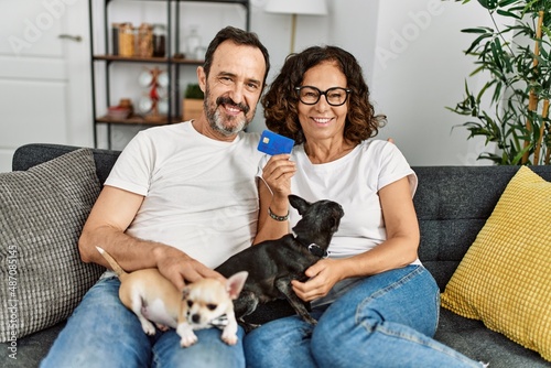 Middle age hispanic couple smiling happy and holding credit card. Sitting on the sofa with dogs at home.