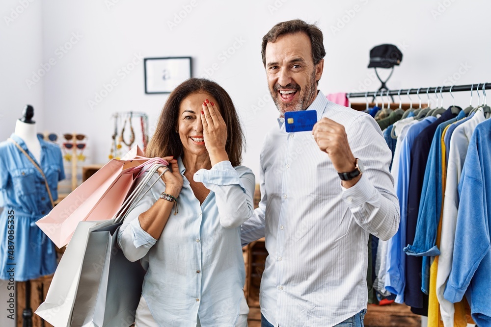 Hispanic middle age couple holding shopping bags and credit card covering one eye with hand, confident smile on face and surprise emotion. Hispanic middle age couple holding shopping bags and credit card covering one eye with hand, confident smile on face and surprise emotion.